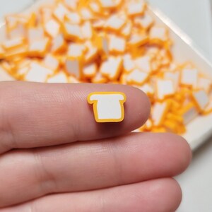 May include: A close-up of a small, white bread charm with a yellow outline. The charm is being held in a hand, and there are many more of the same charms in the background.