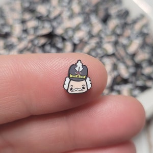 May include: A small, cartoon-style nutcracker figurine with a black hat, white face, and a gold trim. The figurine is being held by a finger.