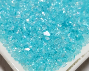 May include: A white ceramic dish filled with iridescent turquoise blue glass glitter. The glitter is small and has a sparkly finish.