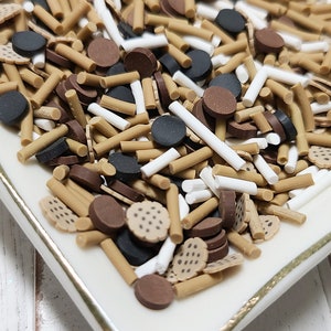 May include: A white ceramic dish with a gold rim filled with brown, white, and black sprinkles. The sprinkles are shaped like sticks, circles, and small squares.