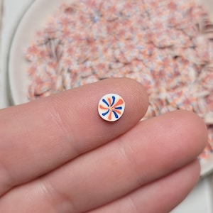 May include: A small, white, circular embellishment with a blue and orange pinwheel design held between fingers. A white ceramic dish filled with similar embellishments is in the background. The embellishments have a floral pattern.