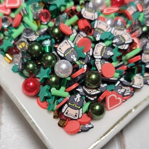 May include: A white plate filled with colorful Christmas sprinkles. The sprinkles are in the shape of stars, hearts, and nutcrackers. There are also red and green beads.