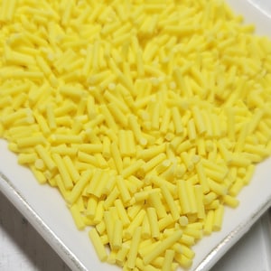 May include: A white square plate filled with yellow sprinkles. The sprinkles are long and thin, resembling small sticks.
