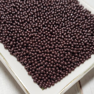 May include: A rectangular white ceramic dish with a gold trim is filled with small, dark brown spherical sprinkles. The sprinkles are densely packed, creating a textured surface. The dish sits on a white, weathered wooden surface.