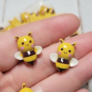 May include: Three yellow and black bee-shaped resin embellishments with white wings and pink cheeks. The bees have a smiling face.