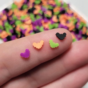 May include: Close-up of four small heart-shaped sprinkles in different colors: purple, orange, green, and black. The sprinkles are on a finger.