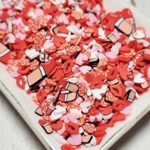 May include: A white ceramic dish filled with red, pink, and white sprinkles. The sprinkles are shaped like hearts, lips, and roses.