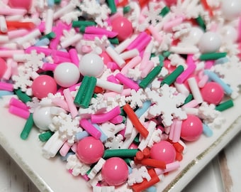 May include: A rectangular white ceramic dish with a gold rim is filled with colorful sprinkles. The sprinkles include pink, white, red, green, and blue sprinkles in various shapes, including small balls, rods, and snowflake shapes.
