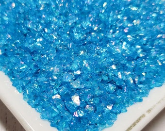 May include: A white ceramic dish filled with iridescent blue glitter. The glitter is small and has a variety of shapes.