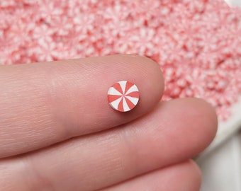 May include: A single red and white candy cane sprinkle, approximately 1/2 inch in diameter, is held between two fingers. The sprinkle is a popular decoration for baking and crafting.