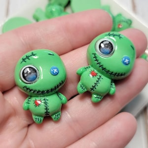 May include: Two green, cartoon-like voodoo doll charms with stitched details, blue accents, and large, expressive eyes. The charms are held in a hand, with other green charms visible in the background. These charms are ideal for jewelry making or crafting.