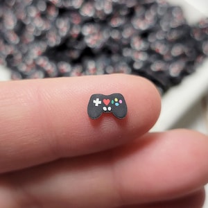 May include: A close-up of a small black and white video game controller charm. The controller has colorful buttons and is held between two fingers.