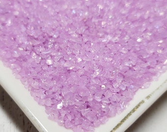 May include: A white ceramic dish filled with iridescent lavender-colored glitter. The glitter is small and has a slightly chunky texture.