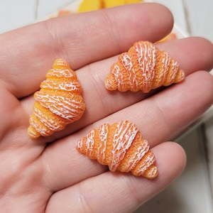 May include: Three miniature orange croissants with white detailing are held in a hand. The croissants are arranged on a white surface with a blurred background of a plate with orange fruit slices. The croissants are a food-themed craft supply.
