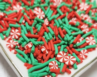 May include: A white square plate with a gold rim filled with red and green candy sprinkles. The sprinkles are in the shape of candy canes and thin sticks.