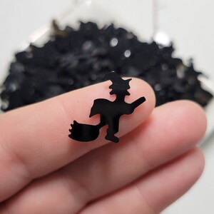 May include: A close-up of a black witch silhouette on a broomstick. The witch is flying to the right. The silhouette is made of a shiny, black material.