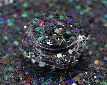 May include: A close-up of a small jar filled with iridescent black glitter. The glitter has a variety of colors, including green, blue, and purple. The jar is sitting on a white surface.