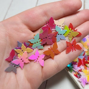 May include: A hand holding a variety of colorful butterfly-shaped sequins. The sequins are iridescent and have a metallic sheen. There are also a number of sequins in a white dish.