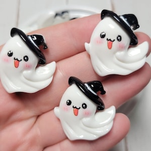 May include: Three white ghost charms with black witch hats and pink cheeks. The ghosts have black eyes and red tongues sticking out.