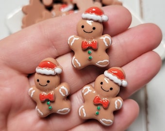 May include: Three small gingerbread man figurines made of smooth, light brown material. Each figure wears a red and white Santa hat, a red bow tie, and has two green dots on the chest. The figures feature white icing-style details on the limbs.