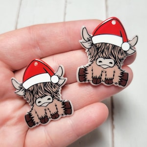 May include: Two small, white ceramic ornaments shaped like Highland cows wearing red Santa hats. The cows are sitting with their legs tucked under them.