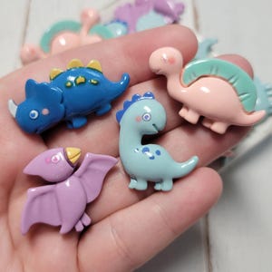May include: A collection of colorful dinosaur figurines in various colors, including blue, pink, and purple. The figurines are small and detailed, with different dinosaur species represented. The dinosaurs are held in a hand, showcasing their size and design.