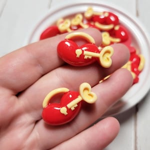 May include: A close-up of a hand holding two red heart-shaped charms with yellow keyhole accents. The charms are in a bowl with other similar charms.
