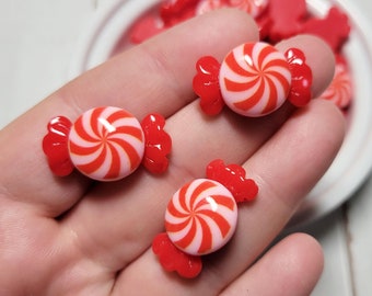 May include: A close-up of three red and white candy cane shaped resin embellishments. The candy canes are on a hand, and there are more candy canes in the background.