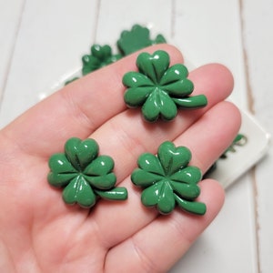 May include: A close-up of three green four-leaf clover charms. The charms are made of a shiny, smooth material and have a slightly raised design.