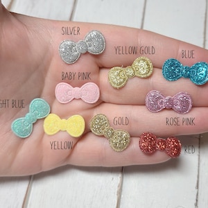 May include: Assortment of glitter bow embellishments in various colors, including silver, yellow gold, blue, baby pink, light blue, yellow, gold, rose pink, and red.