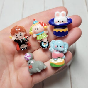 May include: Five colorful figurines of a magician, a clown riding a unicycle, a rabbit in a top hat, an elephant, and a gray mouse with a ball. The figurines are made of resin and have a whimsical, cartoonish style.