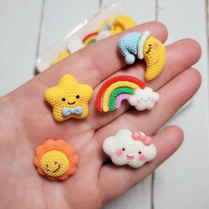May include: A collection of colorful, miniature resin charms in the shape of a smiling star, sun, moon, rainbow, and cloud. The charms are yellow, orange, red, blue, green, and white, with cute facial expressions and details.
