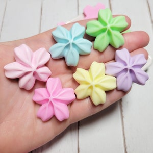 May include: A collection of pastel-colored flower-shaped resin embellishments. The colors include light blue, light green, light pink, lavender, and yellow.