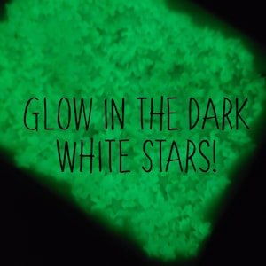 May include: A close-up of a pile of white glow-in-the-dark stars. The stars are glowing green in the dark. The text "GLOW IN THE DARK WHITE STARS!" is superimposed on the image.