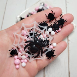 May include: A collection of Halloween-themed craft supplies, including black plastic spiders, pink and white beads, and a white spiderweb. The items are held in a hand, suggesting a small scale and craft project.