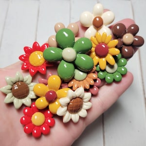 May include: A collection of colorful flower-shaped buttons in various shades of red, green, brown, yellow, and white. The buttons are made of plastic and have a glossy finish.