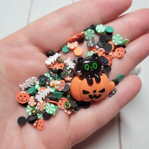 May include: A variety of Halloween-themed sprinkles in a hand, including orange pumpkins, black cats, candy canes, and green leaves.