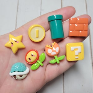 May include: A set of nine colorful resin charms featuring characters and objects from the popular video game Super Mario Bros. The charms include a yellow star, a green pipe, a red brick, a yellow question block, a blue turtle shell, a red flower, a yellow coin, and a pink flower with a red mouth.