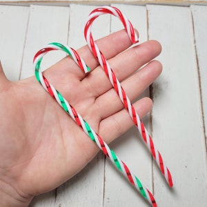May include: Two candy canes, one red and white with green stripes and one red and white with green stripes, are held in a hand.