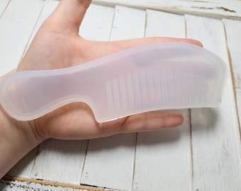 May include: A white silicone comb with a handle and a row of teeth. The comb is held in a hand.