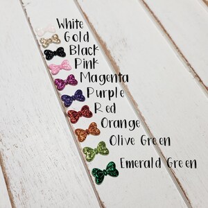 Peut inclure: Une rang&eacute;e de dix n&oelig;uds scintillants de diff&eacute;rentes couleurs, dont le blanc, l'or, le noir, le rose, le magenta, le violet, le rouge, l'orange, le vert olive et le vert &eacute;meraude. Les n&oelig;uds sont dispos&eacute;s verticalement sur une surface en bois blanc.