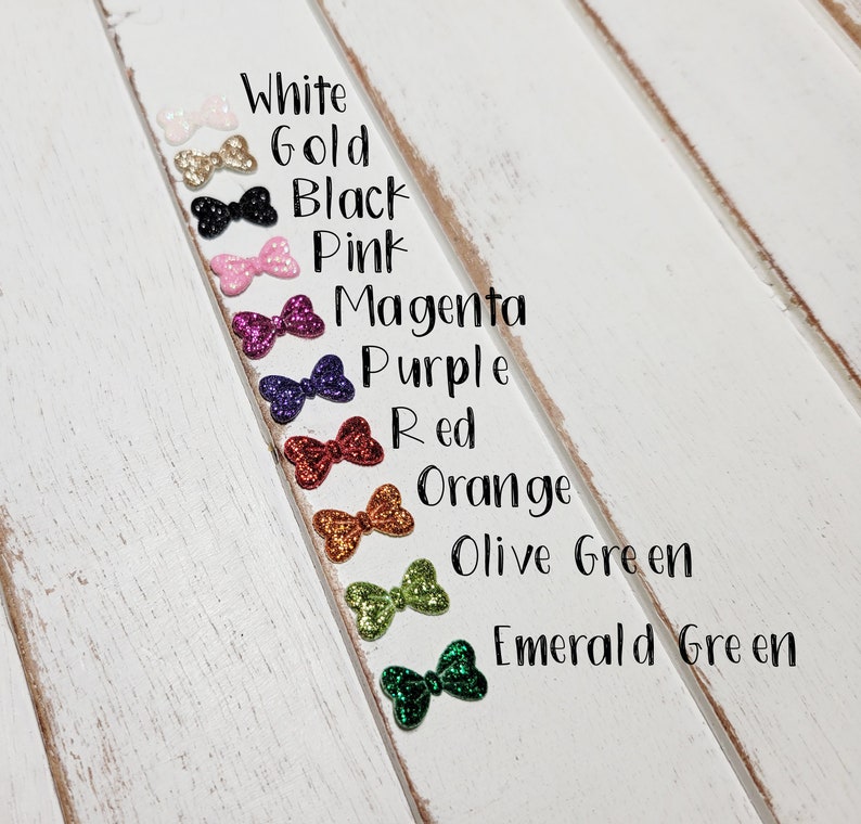 Peut inclure: Une rang&eacute;e de dix n&oelig;uds scintillants de diff&eacute;rentes couleurs, dont le blanc, l'or, le noir, le rose, le magenta, le violet, le rouge, l'orange, le vert olive et le vert &eacute;meraude. Les n&oelig;uds sont dispos&eacute;s verticalement sur une surface en bois blanc.
