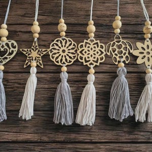 Op de afbeelding: Houten ornamenten met verschillende vormen, waaronder een hart, ster en bloem, elk met een kwastje. De ornamenten zijn opgehangen aan een touwtje en houten kralen. De kwastjes zijn in grijstinten en wit.