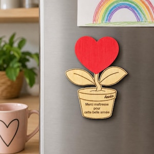 Peut inclure: Un aimant en bois en forme de plante en pot avec un cœur rouge en guise de fleur. Le pot porte l'inscription gravée "Année" et "Merci maîtresse pour cette belle année". Le design comprend deux feuilles en bois clair et une tige fine.