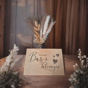 Peut inclure: Une enseigne en bois avec l'inscription "Le petit Bar à Tatouages" et des cœurs. Elle est posée sur une table en bois, avec un bouquet de fleurs séchées en arrière-plan. L'ensemble dégage une atmosphère rustique et chaleureuse, idéale pour un événement.
