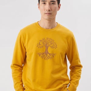 Può includere: Una felpa a girocollo a maniche lunghe color giallo senape, caratterizzata da una grafica centrale a linee marroni raffigurante un albero con radici visibili e una luna crescente integrata tra i rami. Collo, polsini e orlo a costine.