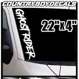 Op de afbeelding: Witte "Ghost Rider" sticker, ongeveer 56 cm x 10 cm, op een zwarte achtergrond, waarschijnlijk een autoraam. De afbeelding toont de tekst "COUNTRYBOYDECALS" en "GHOST RIDER". Extra tekst onderaan.