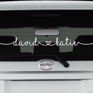 Pode incluir: Adesivo de vinil branco com os nomes "david" e "katie" separados por um símbolo de coração em uma janela de carro preta.