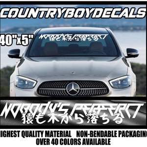 May include: A white car windshield decal with the text "Nobody's Perfect" and Japanese characters. The decal is 40 inches by 5 inches. The decal is on a silver car with a black grill and a Mercedes-Benz logo.