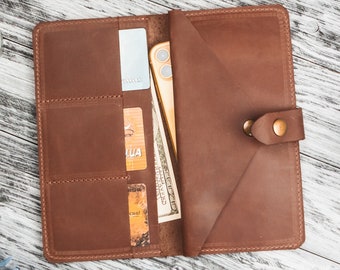 May include: Open brown leather wallet with multiple card slots, a bill compartment, and a snap closure. The wallet contains a smartphone, credit cards, and cash. The wallet is on a wooden surface.